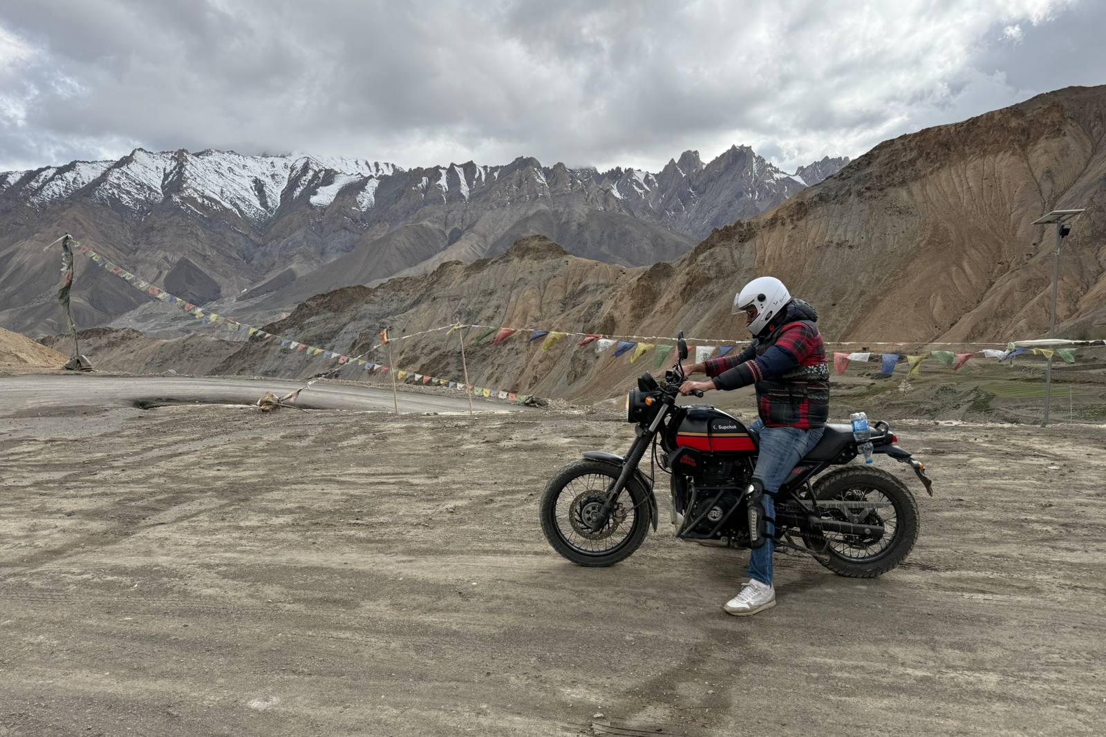 Motociclista en una Royal Enfield Himalayan junto a banderas de oración y montañas nevadas, cerca de Lamayuru, en Ladakh