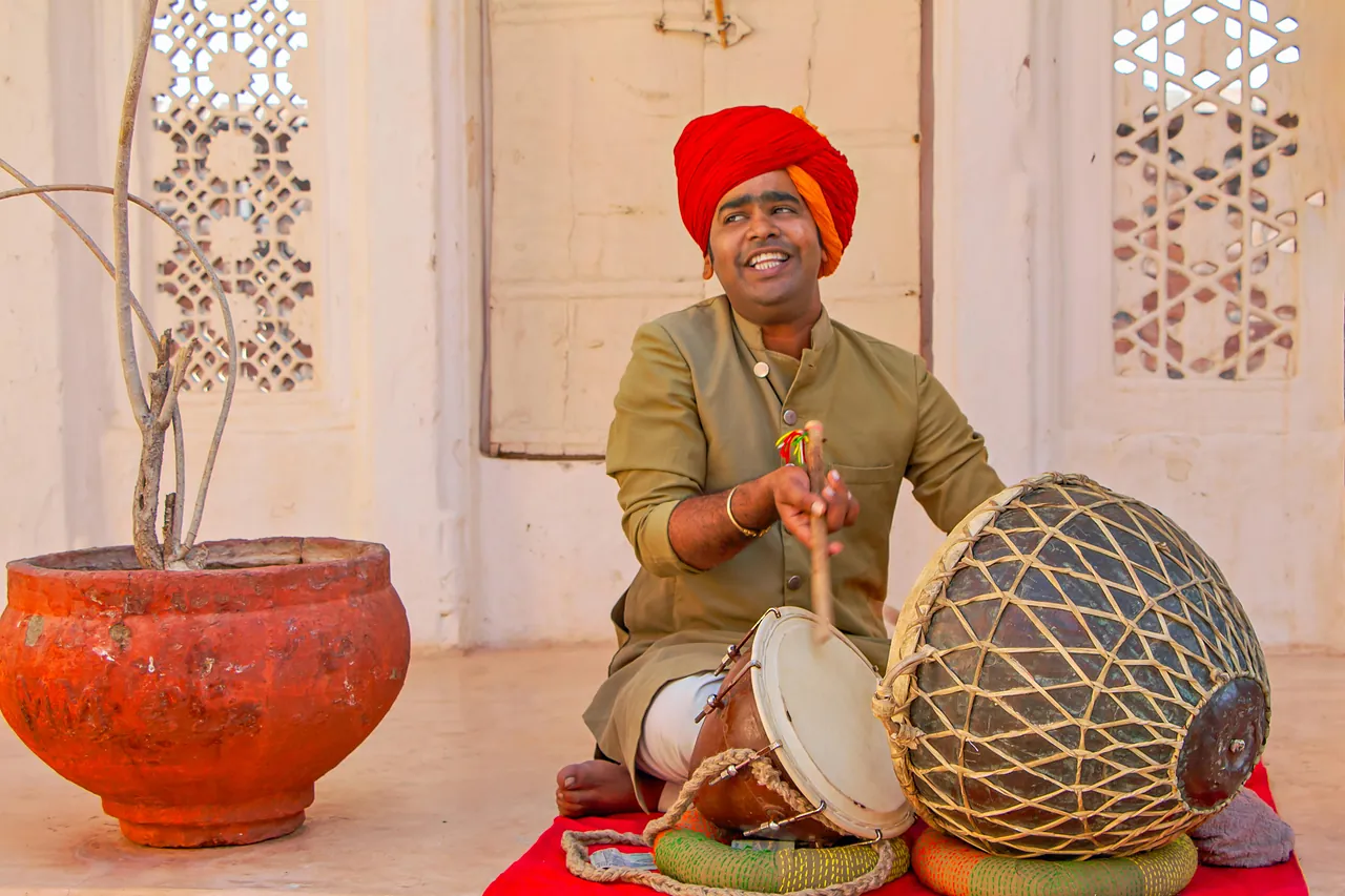 Músico rajastaní con turbante rojo tocando el dholak junto a una celosía