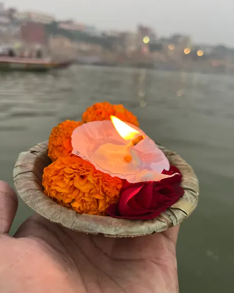 Una mano sostiene un cuenco de hojas con una vela encendida, caléndulas y una rosa, antes de ofrecerlo al Ganges en Varanasi