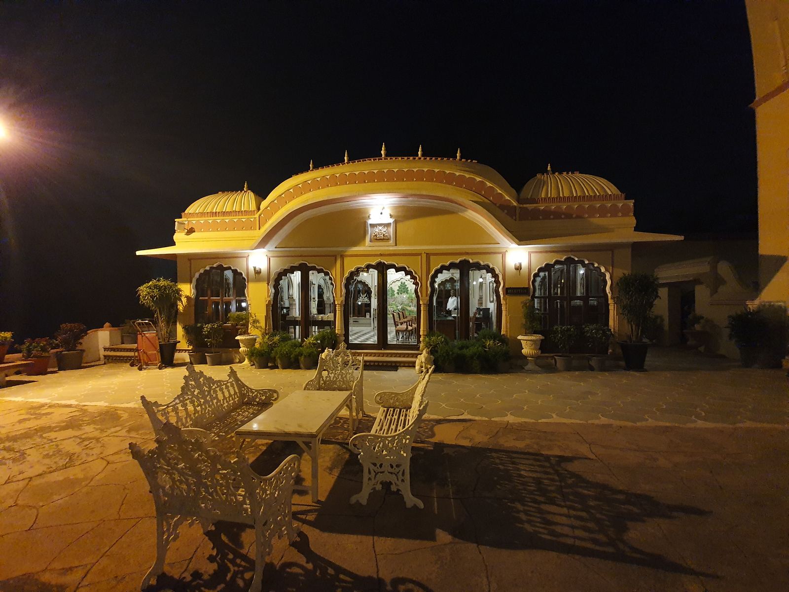 Fachada de una casa señorial india iluminada de noche, con arcos lobulados y una terraza con sillones de forja